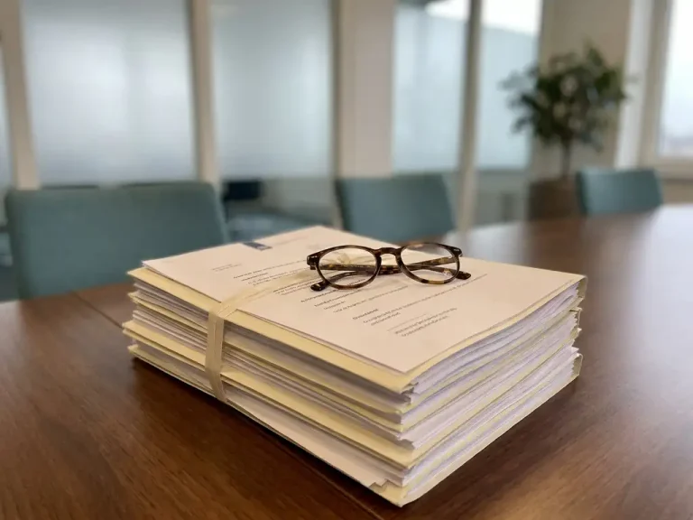 Stapel Nederlandse zorgdocumenten en arbeidscontracten op vergadertafel met leesbril, zacht ochtendlicht door matglazen ramen.